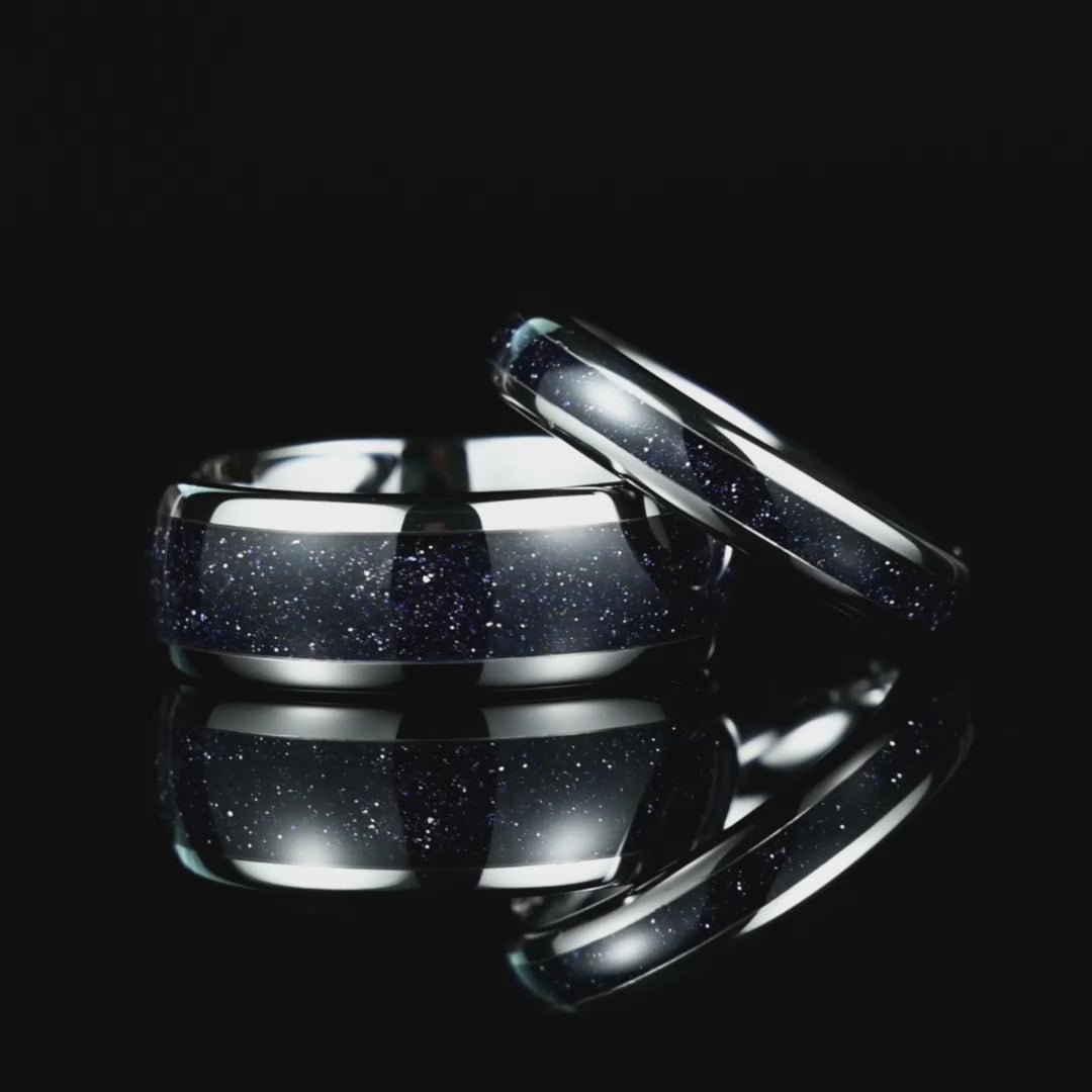 A video of his and her silver tungsten wedding rings with galaxy glow design rotating on a reflective black glass surface, highlighting their cosmic pattern.